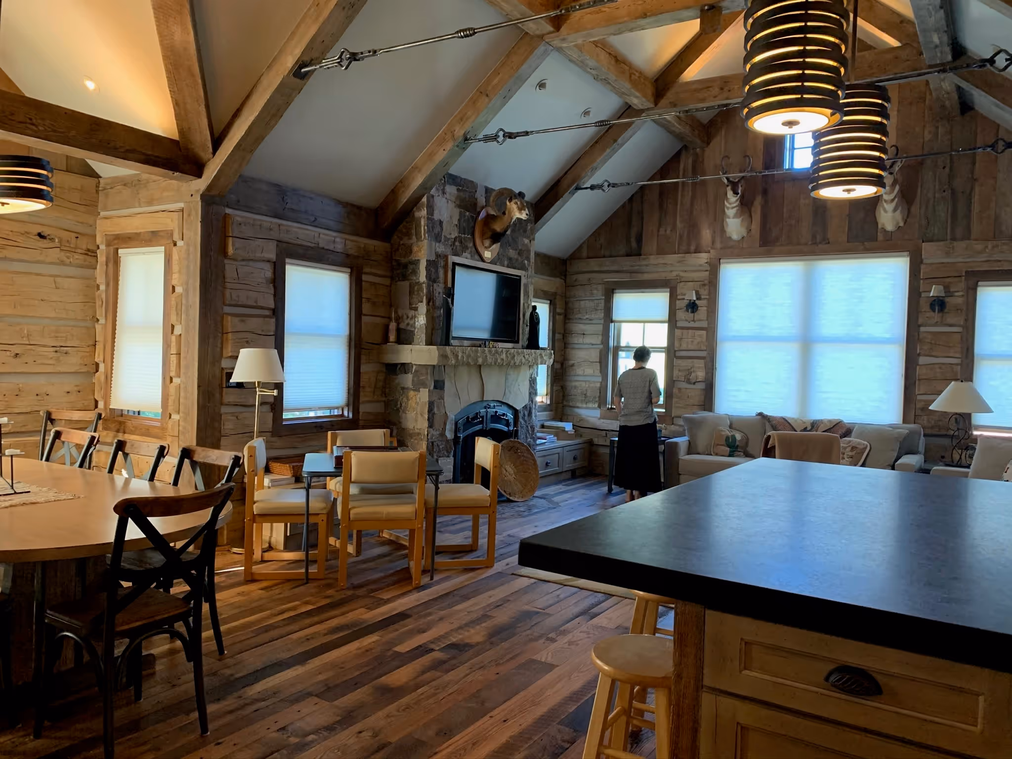 A rustic living room with exposed wooden beams, stone fireplace, and large windows letting in natural light. The room is furnished with a dining table, chairs, couches, and lamps, while a person stands near the back by a window.