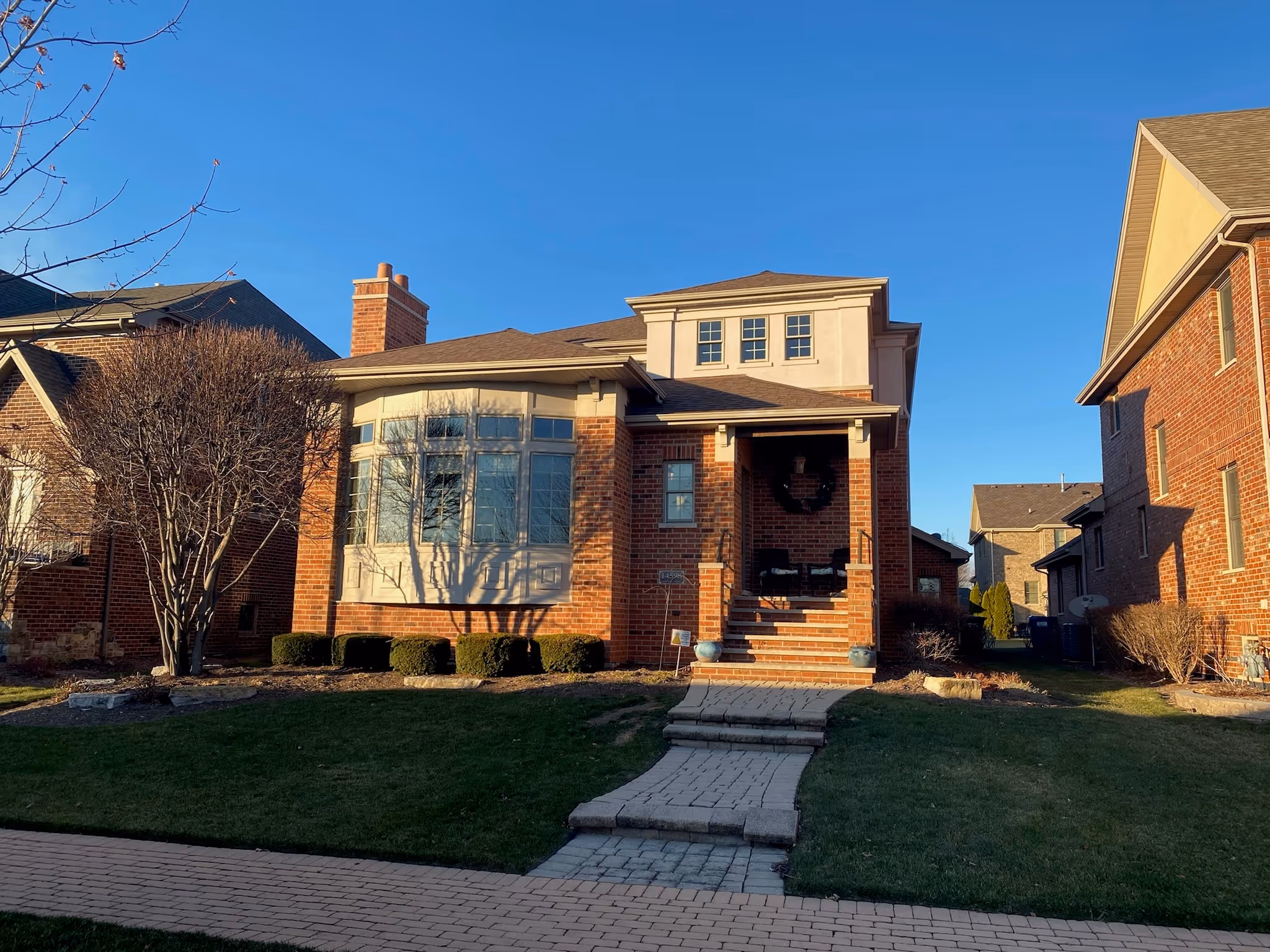 Red brick house with a front porch, bay window, shrubs, and a paved walkway on a clear day.