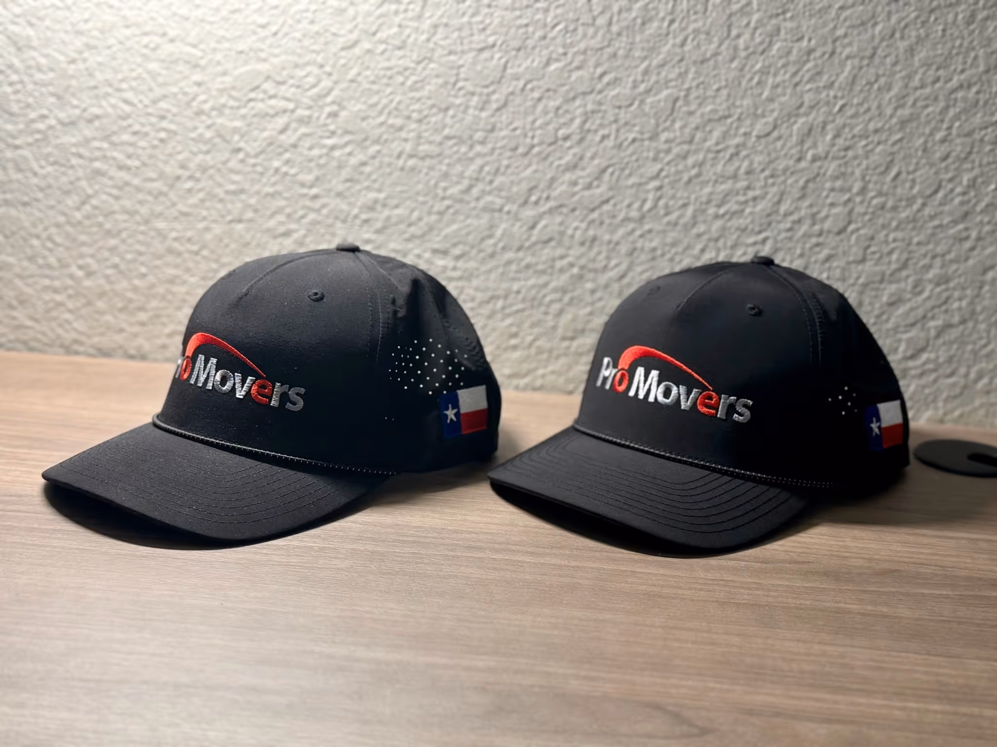 Two black ProMovers caps with Texas flags on a wood surface against a textured wall.