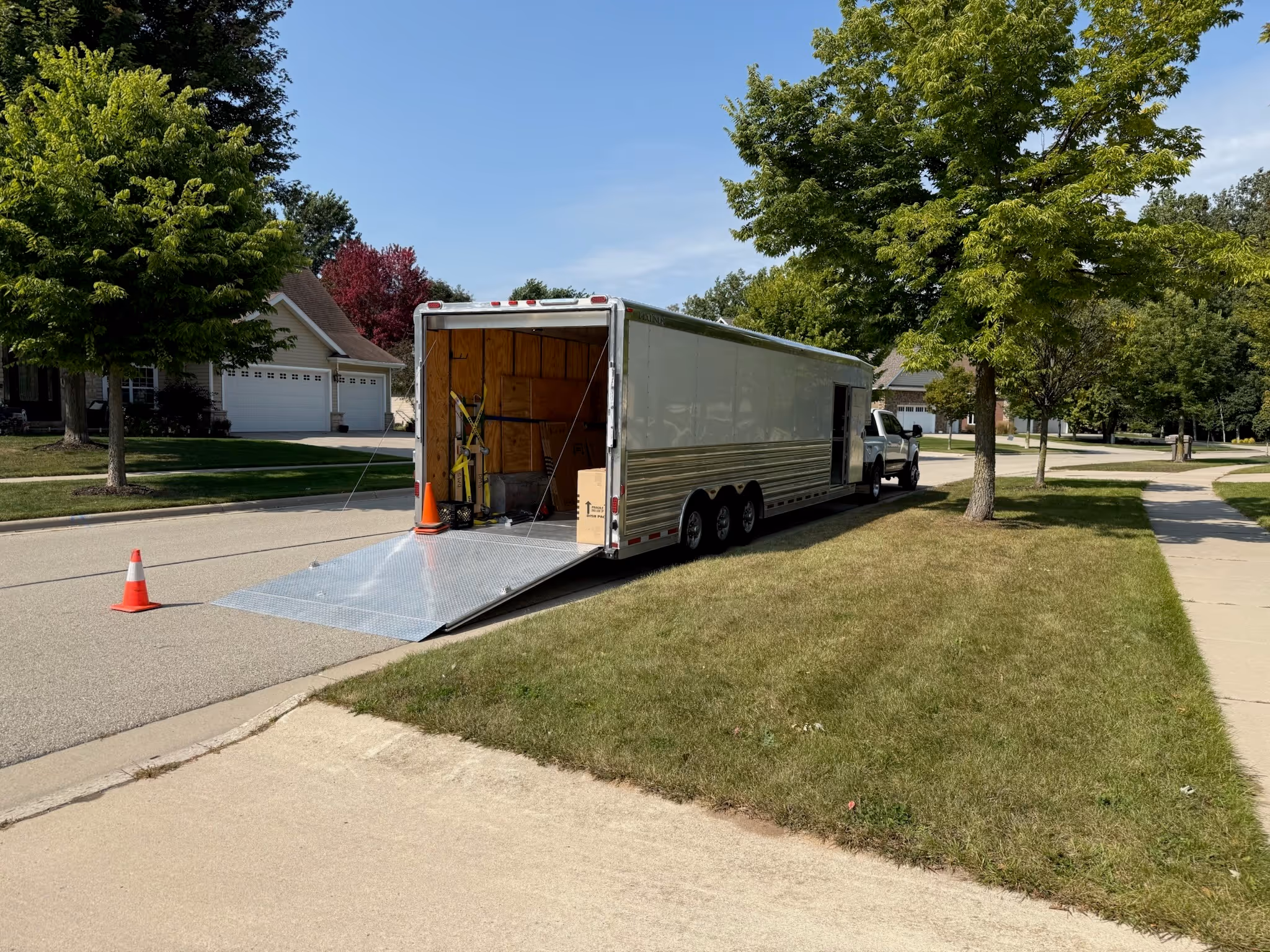 A large enclosed moving trailer with its rear ramp down is parked on a suburban street beside a grassy lawn, with orange cones placed near the ramp for safety.