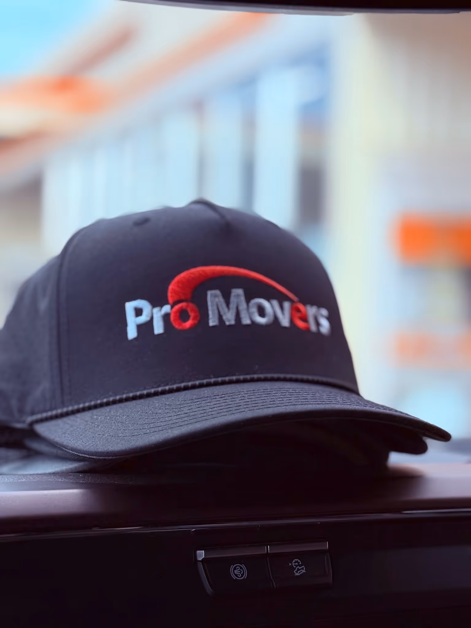 A black baseball cap with the embroidered logo “Pro Movers” in white and red lettering rests on a car dashboard.