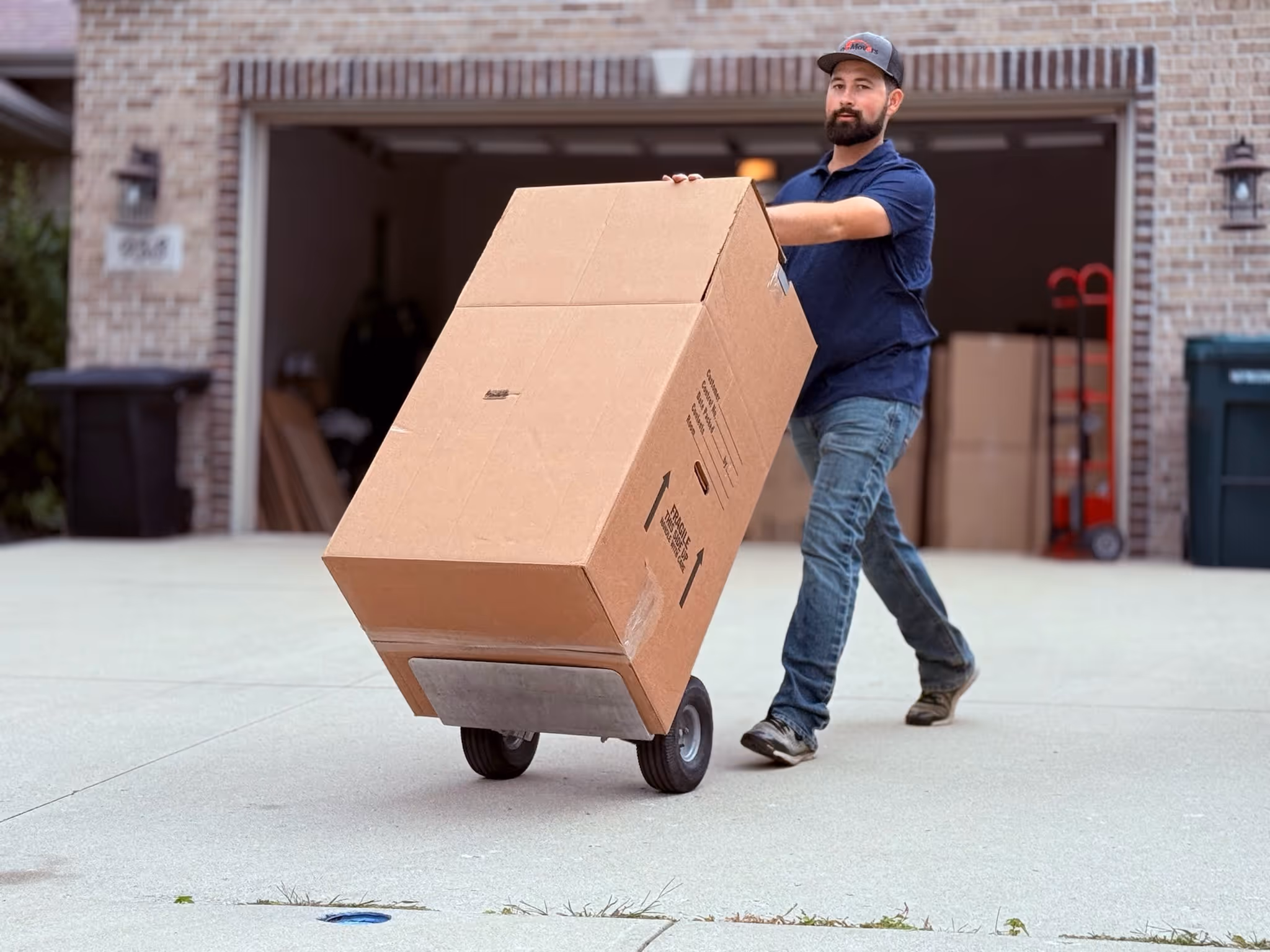 A man wearing a navy blue shirt, jeans, and a cap is using a hand truck to move a large cardboard box up a driveway in front of an open garage.