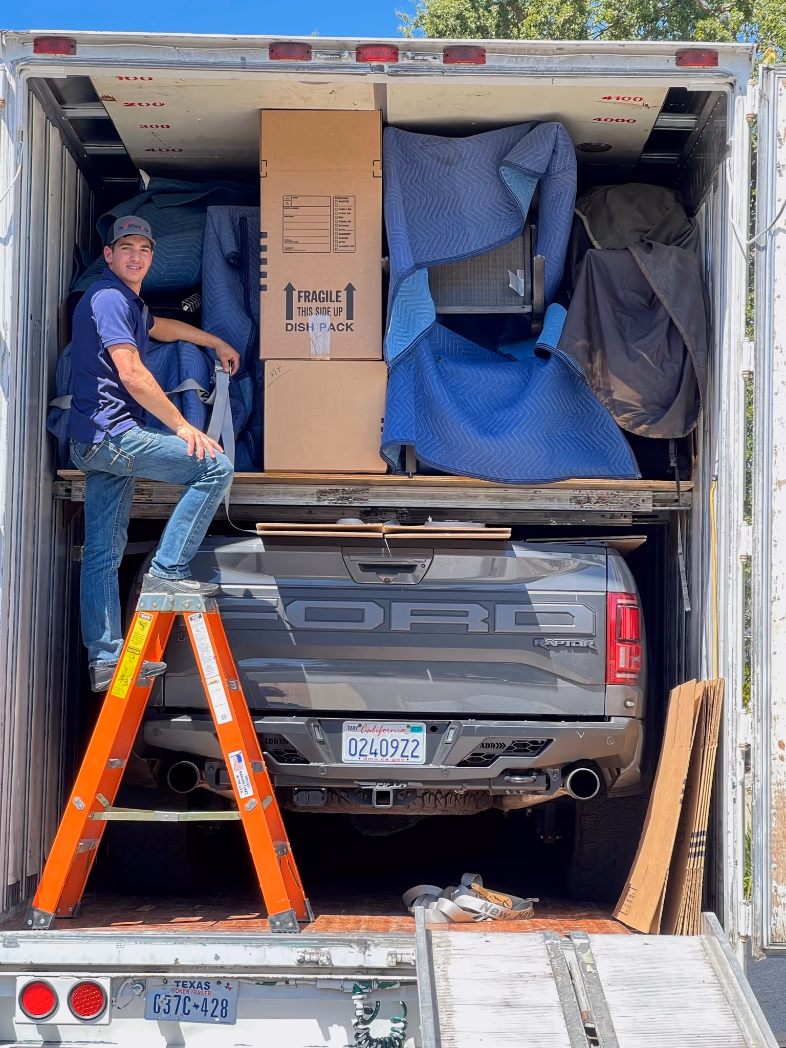 A mover stands on an orange ladder inside a truck, organizing large boxes and furniture wrapped in moving blankets stacked above the bed of a gray Ford pickup being transported.